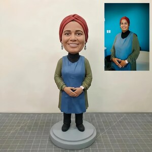 Puede incluir: Una figura de bobblehead personalizada de una mujer que lleva un tocado rojo, un cuello alto negro, un delantal azul y una camisa verde de manga larga. La figura está de pie sobre una base circular gris. Una imagen más pequeña de la mujer está en el fondo.