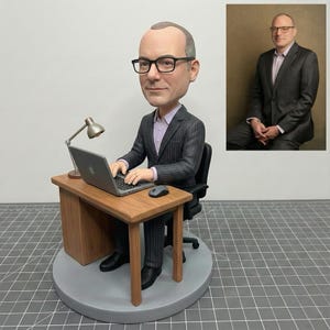 Könnte beinhalten: Eine individuelle Wackelkopf-Figur eines Mannes mit Brille, Anzug und lila Hemd, der an einem Schreibtisch mit Laptop, Maus und Lampe sitzt. Ein Foto des Mannes im Anzug befindet sich im Hintergrund.