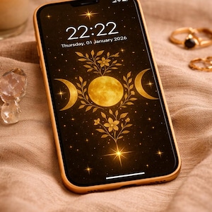 May include: A gold-colored smartphone displays a celestial design with a full moon, crescent moons, and stars on a black background. The time 22:22 and the date Thursday, 01 January 2026 are visible. Gold rings and a candle are in the background.