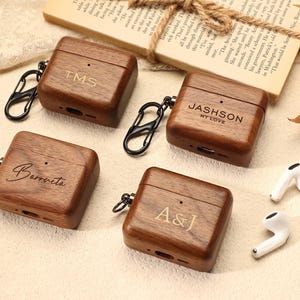 May include: Four wooden AirPod cases with black carabiner clips, each engraved with initials or names: "TMS", "Barrveta", "A&J", and "JASHSON MY LOVE". Two white AirPod earbuds are also visible. The cases are made of wood.