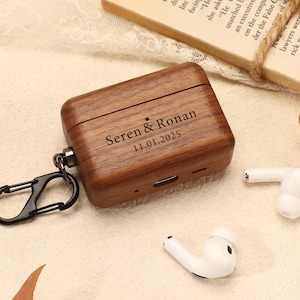 Könnte beinhalten: Eine braune Holz-AirPod-Hülle mit den eingravierten Namen "Seren & Ronan" und dem Datum "11.01.2025". Sie hat einen schwarzen Karabinerhaken. Zwei weiße AirPods liegen daneben.
