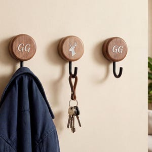 May include: Three wooden wall hooks with black metal hooks. Two hooks have the initials "GG" in a silver color, and one has a deer silhouette. A blue jacket hangs on one hook, and keys hang from another.