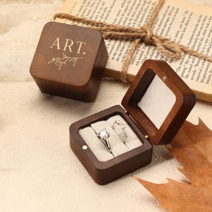 May include: Two wooden ring boxes, one open and one closed, sit on a cream-colored surface. The open box displays two rings on a white cushion. The closed box has the word "ART." and a leaf design engraved on the lid. A book and leaves are in the background.