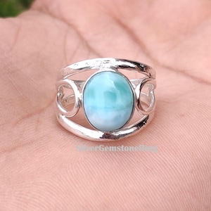 May include: A silver ring featuring a large, oval-shaped, light blue gemstone. The ring has a decorative design on the sides. The gemstone is the focal point, showcasing a blend of blue and white hues. The ring is a piece of jewelry.