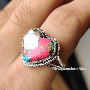 Puede incluir: Anillo de plata con una piedra preciosa en forma de corazón. La piedra preciosa tiene una mezcla de colores, incluyendo rosa, blanco y turquesa. El anillo tiene un borde trenzado y un diseño de caña dividida. El texto "SilverGemstoneRing" es visible.
