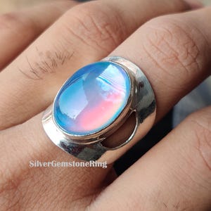 May include: A silver ring with an oval, iridescent gemstone. The gemstone transitions from blue to pink, set in a wide, decorative band. The ring is shown on a hand.
