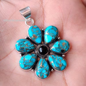 May include: A silver pendant with a flower design. The pendant features turquoise teardrop-shaped stones with copper-colored veining, surrounding a black circular stone. The pendant is approximately 1.5 inches in diameter.