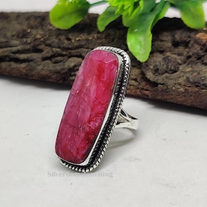 May include: A silver ring featuring a large, rectangular, ruby-colored gemstone. The gemstone is set in a silver band with a textured border. The ring is displayed on a light surface with a wooden background and green foliage.