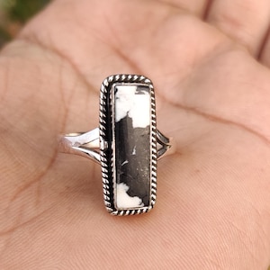 Puede incluir: Un anillo de plata con una piedra rectangular orientada verticalmente con un patrón de mármol blanco y negro. La piedra está engastada en un borde de metal trenzado y la banda tiene un diseño dividido en los lados.