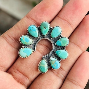 Puede incluir: Un broche de plata en forma de herradura con ocho piedras de turquesa en forma de lágrima. El marco de plata tiene una textura de cuerda trenzada en la curva interior, y cada piedra está engastada individualmente en un soporte de plata pulida.