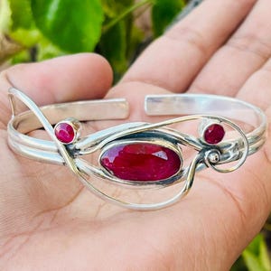 Puede incluir: Brazalete de plata con un gran rubí ovalado y dos rubíes redondos más pequeños. El diseño abierto presenta remolinos decorativos. El brazalete muestra las vibrantes piedras rojas. La circunferencia interior del brazalete es de unos 15 cm.