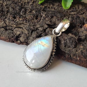 May include: A teardrop-shaped moonstone pendant with a silver bail. The iridescent moonstone is set in a silver bezel with a rope-like border. The pendant is displayed on a dark, textured surface.