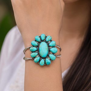May include: A silver cuff bracelet featuring a large central oval turquoise stone surrounded by twelve smaller pear-shaped turquoise stones arranged in a floral pattern. The metal band is split into two thin silver wires on each side of the setting.