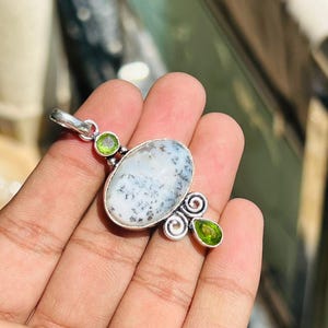 May include: A silver pendant featuring an oval, white gemstone with black inclusions. Two green gemstones and decorative swirls accent the pendant. The pendant is held in a hand.