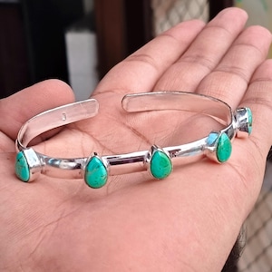Könnte beinhalten: Ein silbernes Armband mit vier tropfenförmigen Türkissteinen, die entlang des äußeren Bandes eingefasst sind. Die Metalloberfläche ist poliert und reflektierend, mit einem 925er Stempel auf der Innenseite des offenen Armreifdesigns.