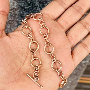 Peut inclure: Un bracelet de couleur or rose composé d'une série de maillons circulaires interconnectés de tailles variées. Le bracelet est fixé par un mécanisme de fermoir à barre en T, constitué d'une barre métallique horizontale glissant dans un anneau.