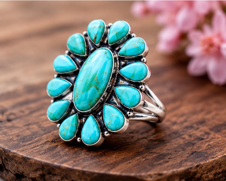 Puede incluir: Un anillo de plata con una gran piedra turquesa ovalada en el centro, rodeada por doce piedras turquesas m&aacute;s peque&ntilde;as en forma de pera dispuestas en un patr&oacute;n floral. El engaste de metal tiene un acabado pulido con detalles de cuentas.