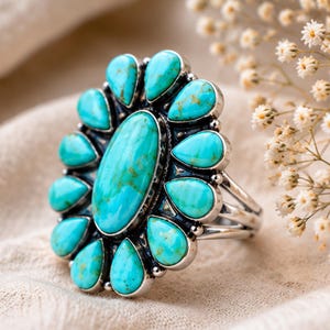 Puede incluir: Un anillo de plata con una piedra turquesa grande de forma ovalada en el centro, rodeada por doce piedras turquesas m&aacute;s peque&ntilde;as en forma de l&aacute;grima dispuestas en un patr&oacute;n floral. El engaste de metal tiene un acabado pulido con oxidaci&oacute;n oscura.