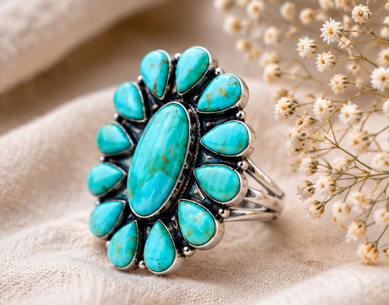 Puede incluir: Un anillo de plata con una piedra turquesa grande de forma ovalada en el centro, rodeada por doce piedras turquesas m&aacute;s peque&ntilde;as en forma de l&aacute;grima dispuestas en un patr&oacute;n floral. El engaste de metal tiene un acabado pulido con oxidaci&oacute;n oscura.