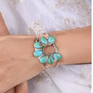 K&ouml;nnte beinhalten: Ein silbernes Armband mit einem zentralen hufeisenf&ouml;rmigen Metallrahmen, der mit sieben tropfenf&ouml;rmigen T&uuml;rkissteinen verziert ist. Die hellblauen Steine weisen braune Aderungen auf und sind f&auml;cherf&ouml;rmig um die Mitte angeordnet.