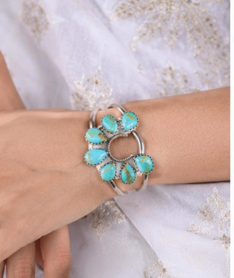 K&ouml;nnte beinhalten: Ein silbernes Armband mit einem zentralen hufeisenf&ouml;rmigen Metallrahmen, der mit sieben tropfenf&ouml;rmigen T&uuml;rkissteinen verziert ist. Die hellblauen Steine weisen braune Aderungen auf und sind f&auml;cherf&ouml;rmig um die Mitte angeordnet.