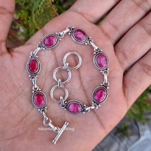 Könnte beinhalten: Ein silbernes Armband mit ovalen, tiefroten Edelsteinen. Das Armband hat ein dekoratives, gedrehtes Seildesign um jeden Stein und einen Knebelverschluss. Der Text "Silver Gems Store Ring" ist sichtbar.