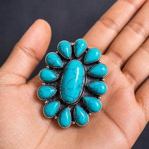 Puede incluir: Un anillo grande con una piedra turquesa ovalada central rodeada por doce piedras turquesas m&aacute;s peque&ntilde;as en forma de l&aacute;grima. Las piedras est&aacute;n engastadas en un marco de plata oxidada oscura con un dise&ntilde;o de borde texturizado.