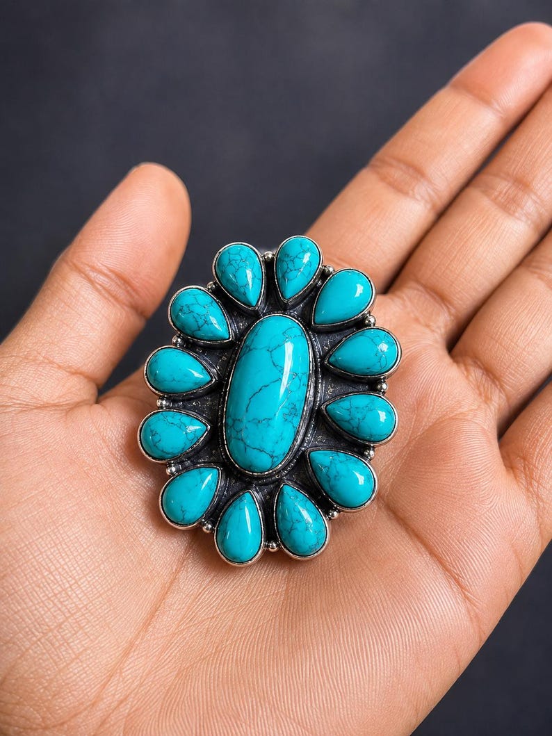 Puede incluir: Un anillo grande con una piedra turquesa ovalada central rodeada por doce piedras turquesas m&aacute;s peque&ntilde;as en forma de l&aacute;grima. Las piedras est&aacute;n engastadas en un marco de plata oxidada oscura con un dise&ntilde;o de borde texturizado.