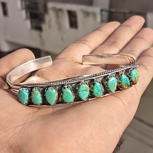 May include: A silver cuff bracelet featuring a row of nine teardrop-shaped turquoise stones. Each stone is set in a scalloped silver bezel, and the band is accented with a twisted silver wire detail along the top edge of the setting.