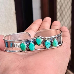 Könnte beinhalten: Ein silberner Armreif mit fünf tropfenförmigen Türkissteinen, die in einer Reihe auf der Vorderseite angeordnet sind. Das Metallband hat eine polierte, reflektierende Oberfläche mit einer Öffnung auf der Rückseite und zeigt die Steine.