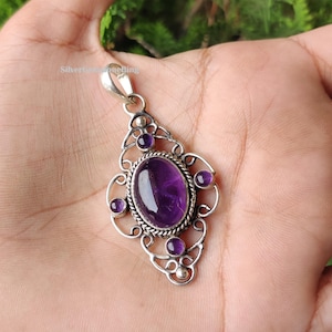 May include: A silver pendant with an oval, deep purple amethyst gemstone at the center, surrounded by smaller purple stones and an ornate, openwork design. The pendant has a loop at the top for a chain.
