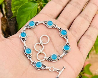 May include: Silver bracelet with light blue, round gemstones set in decorative bezels. The bracelet, with a toggle clasp, is shown on a hand, highlighting the jewellery's design and colour.