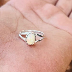 May include: A silver ring with an oval, iridescent opal gemstone. The ring has a split shank design, with two bands that curve around the finger. The opal has a milky white color with flashes of color.
