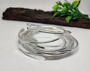 Puede incluir: Un conjunto de siete pulseras de plata apiladas unas sobre otras. Las pulseras tienen todas el mismo ancho y un acabado liso y pulido.