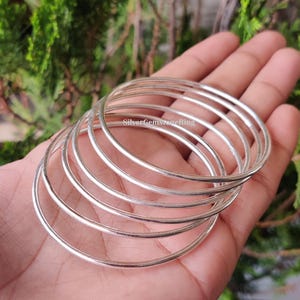 May include: A collection of silver bangles, each a simple, smooth band. The bangles are displayed on a hand, with the text "SilverGemstoneRing" visible. The bangles are made of a shiny, reflective metal.