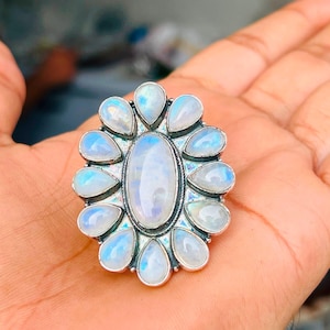 Puede incluir: Un anillo de plata con una gran piedra lunar ovalada central rodeada por doce piedras lunares m&aacute;s peque&ntilde;as en forma de pera dispuestas en un patr&oacute;n floral. Las piedras muestran un aspecto blanco transl&uacute;cido con iridiscencia azul.