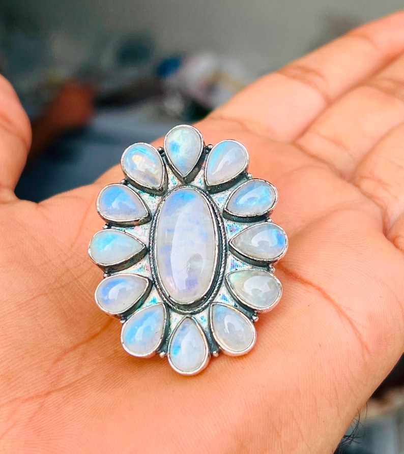 Puede incluir: Un anillo de plata con una gran piedra lunar ovalada central rodeada por doce piedras lunares m&aacute;s peque&ntilde;as en forma de pera dispuestas en un patr&oacute;n floral. Las piedras muestran un aspecto blanco transl&uacute;cido con iridiscencia azul.