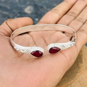 Può includere: Un bracciale a polsino color argento con pietre preziose rosse a forma di goccia a ciascuna estremità. Il bracciale presenta dettagli decorativi vicino alle pietre preziose.