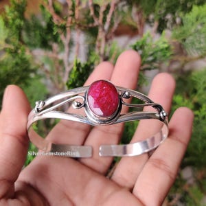 May include: A silver cuff bracelet with a large, oval ruby gemstone. The ruby is a vibrant red colour and is the focal point. The bracelet has an open design with decorative swirls. The text "SilverGemstoneRing" is visible.