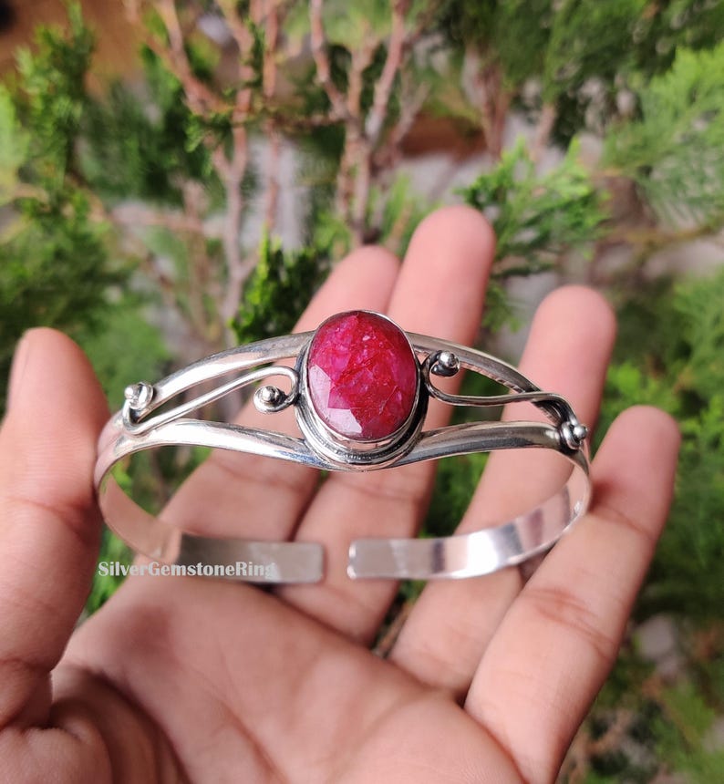 May include: A silver cuff bracelet with a large, oval ruby gemstone. The ruby is a vibrant red colour and is the focal point. The bracelet has an open design with decorative swirls. The text "SilverGemstoneRing" is visible.