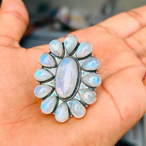 Puede incluir: Un anillo de plata con una gran piedra lunar ovalada central rodeada por doce piedras lunares m&aacute;s peque&ntilde;as en forma de l&aacute;grima. Las piedras presentan un aspecto blanco lechoso y transl&uacute;cido con una sutil iridiscencia azul.