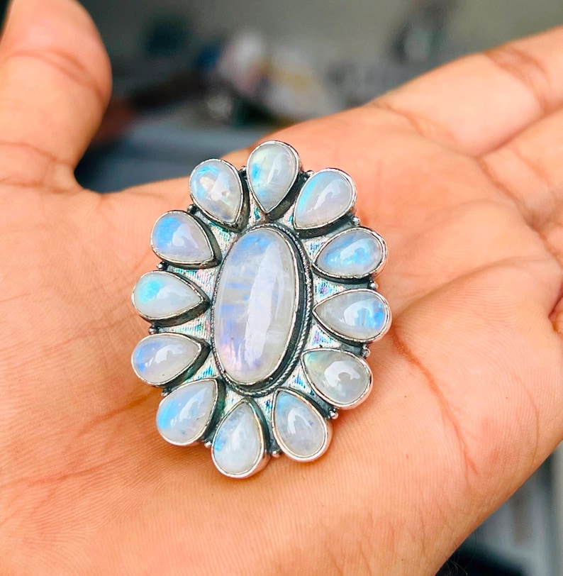 Puede incluir: Un anillo de plata con una gran piedra lunar ovalada central rodeada por doce piedras lunares m&aacute;s peque&ntilde;as en forma de l&aacute;grima. Las piedras presentan un aspecto blanco lechoso y transl&uacute;cido con una sutil iridiscencia azul.