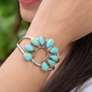 K&ouml;nnte beinhalten: Ein silbernes Armband mit einem zentralen hufeisenf&ouml;rmigen Design, das mit neun tropfenf&ouml;rmigen T&uuml;rkissteinen verziert ist. Die Steine zeigen eine leuchtend blaue Farbe mit brauner Matrix-Maserung und sind jeweils in einer gezackten Silberfassung gefasst.