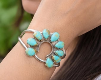 May include: A silver cuff bracelet featuring a central horseshoe-shaped design adorned with nine teardrop-shaped turquoise stones. The stones display a bright blue colour with brown matrix veining, each set in a scalloped silver bezel setting.