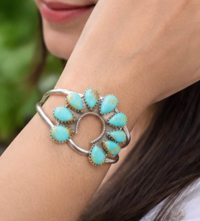 K&ouml;nnte beinhalten: Ein silbernes Armband mit einem zentralen hufeisenf&ouml;rmigen Design, das mit neun tropfenf&ouml;rmigen T&uuml;rkissteinen verziert ist. Die Steine zeigen eine leuchtend blaue Farbe mit brauner Matrix-Maserung und sind jeweils in einer gezackten Silberfassung gefasst.