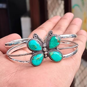 May include: A silver cuff bracelet featuring a butterfly-shaped centrepiece adorned with four teardrop-shaped turquoise stones. The metal band includes a twisted wire detail, and the butterfly wings are outlined with a beaded silver texture.