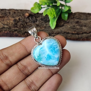 Puede incluir: Un colgante de plata en forma de coraz&oacute;n con una piedra preciosa azul y blanca. El colgante est&aacute; engastado en un marco de plata con un borde decorativo. El colgante est&aacute; en una mano.