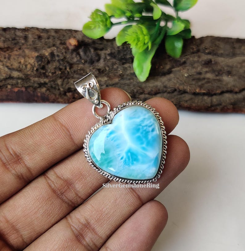 Puede incluir: Un colgante de plata en forma de coraz&oacute;n con una piedra preciosa azul y blanca. El colgante est&aacute; engastado en un marco de plata con un borde decorativo. El colgante est&aacute; en una mano.
