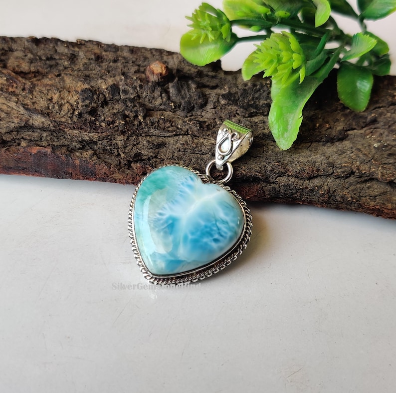 Puede incluir: Un colgante en forma de coraz&oacute;n con una piedra preciosa azul y blanca engastada en plata de ley. El colgante tiene un delicado borde de cadena de plata.