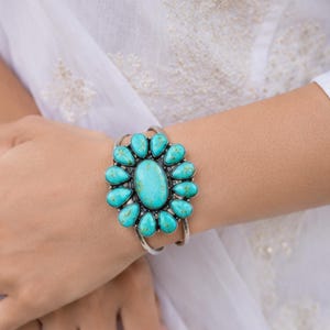 Könnte beinhalten: Ein silbernes Armband mit einem großen, ovalen, zentralen Türkisstein, der von zwölf kleineren, tropfenförmigen Türkissteinen in einem Blumenmuster umgeben ist. Die Metallfassung hat eine polierte Oberfläche und ein geteiltes Armbanddesign.